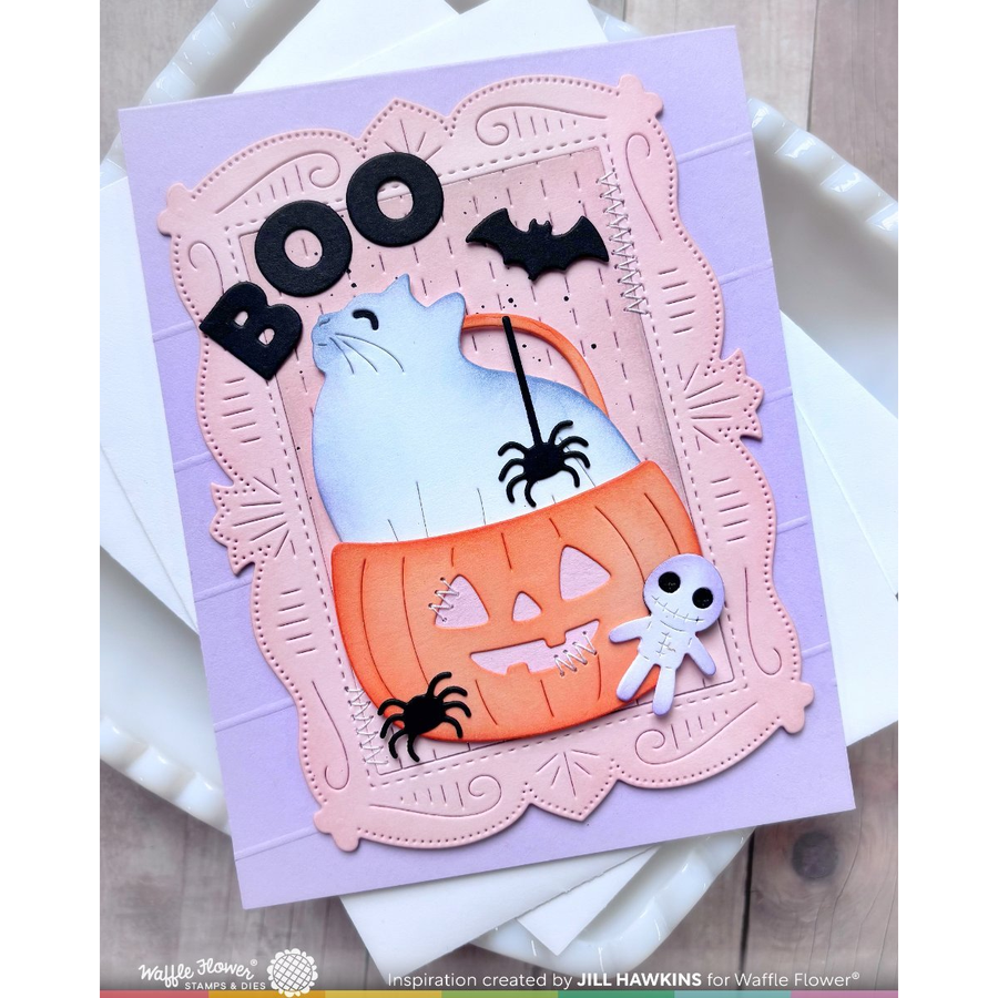 A handmade Halloween greeting card featuring a white cat sitting in an orange jack-o'-lantern bucket, surrounded by a scalloped pink frame. The card includes black 'BOO' lettering, black bats, black spiders, a small white skeleton, and a lavender background, photographed on white envelopes over a light wood surface.