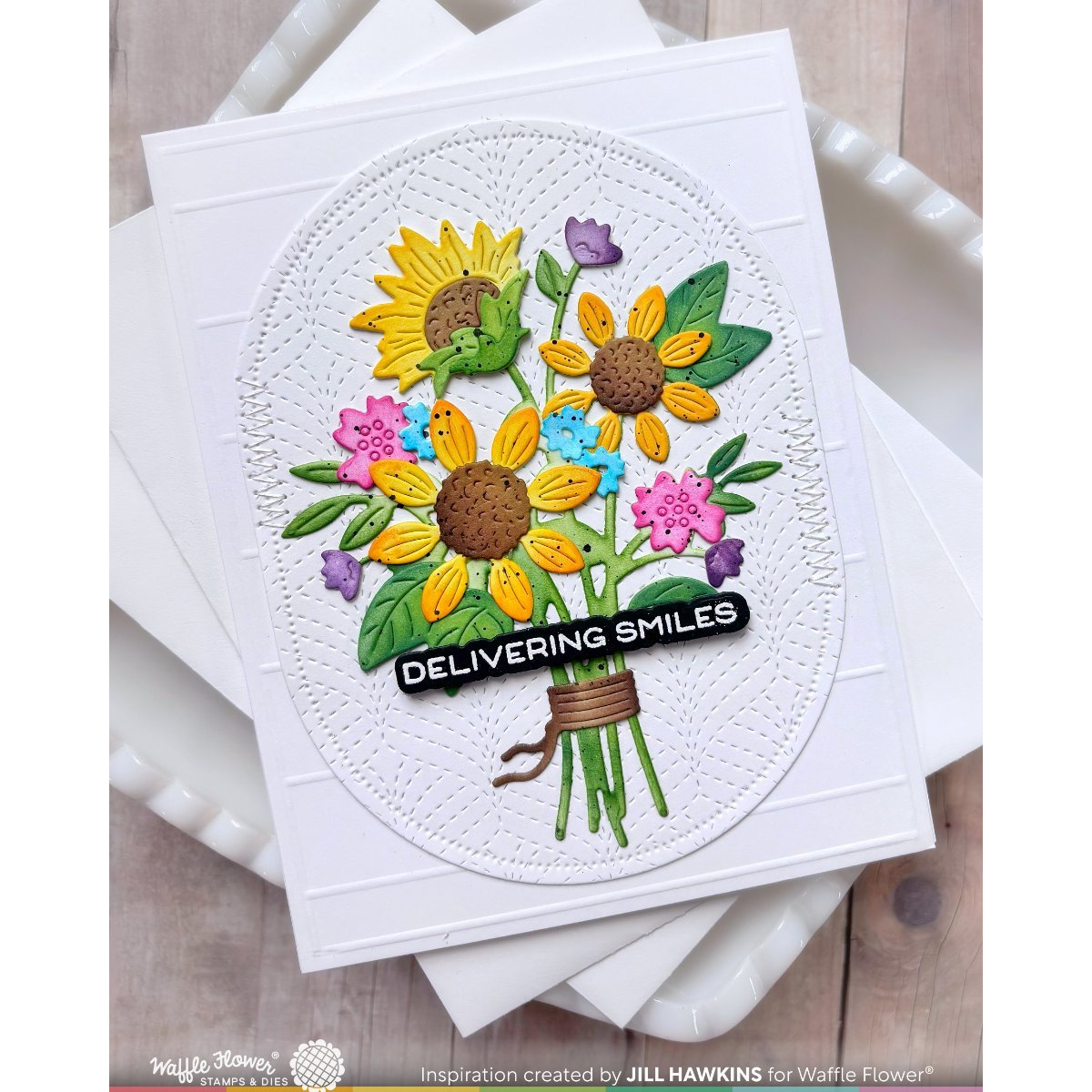A handmade greeting card featuring a 3D floral bouquet with sunflowers and pink, blue, and purple flowers, wrapped in a black banner that reads 'DELIVERING SMILES'. The card is displayed on top of other white cards over a light wood surface.