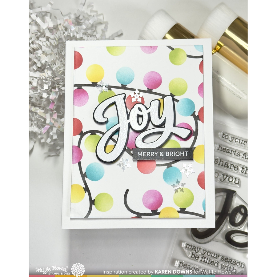 A handmade greeting card featuring a white background decorated with colorful balloon lights in pink, yellow, green, blue, and purple. The word 'Joy' is written in large white cursive script with a black outline, and a black banner below reads 'MERRY & BRIGHT'.