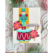 A handmade Christmas card featuring a nutcracker standing on a drum with pink poinsettia flowers and the text 'DRUMROLL FOR CHRISTMAS,' styled on a rustic wood surface with holiday greenery, scissors, candy canes, and white snowflake decorations.