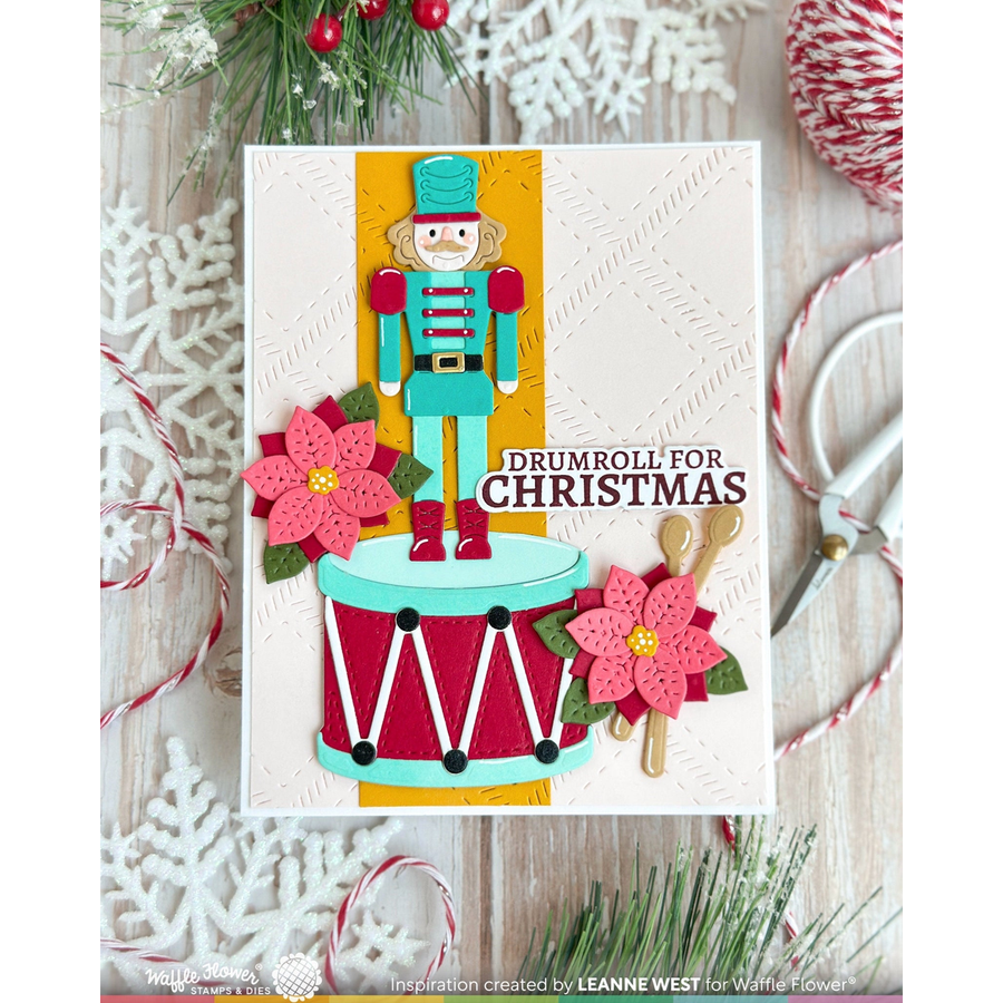 A handmade Christmas card featuring a nutcracker standing on a drum with pink poinsettia flowers and the text 'DRUMROLL FOR CHRISTMAS,' styled on a rustic wood surface with holiday greenery, scissors, candy canes, and white snowflake decorations.