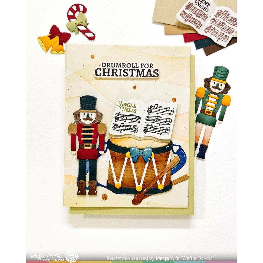 A Christmas-themed card featuring a drum player nutcracker and sheet music, with the text 'DRUMROLL FOR CHRISTMAS.' The card is shown on a white background with small decorative holiday props around it, including bells, candy canes, bows, and additional music-note elements.