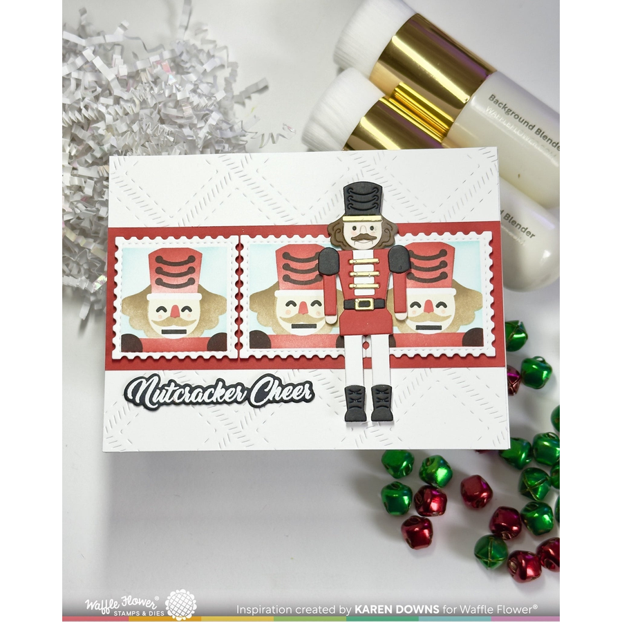 A handmade Christmas nutcracker-themed greeting card displayed on a white surface with silver tinsel, markers, and red and green jingle bells. The card features three nutcracker illustrations in a stamp-like border and the text 'Nutcracker Cheer'.
