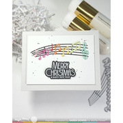 A handmade greeting card with a white background, featuring a colorful musical note and staff line above the words 'MERRY CHRISTMAS & HAPPY NEW YEAR' in black and white. The card is styled with silver tinsel, white and gold cosmetic bottles, and metal dies on a white surface.