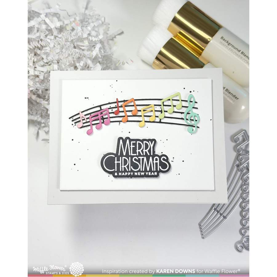 A handmade greeting card with a white background, featuring a colorful musical note and staff line above the words 'MERRY CHRISTMAS & HAPPY NEW YEAR' in black and white. The card is styled with silver tinsel, white and gold cosmetic bottles, and metal dies on a white surface.