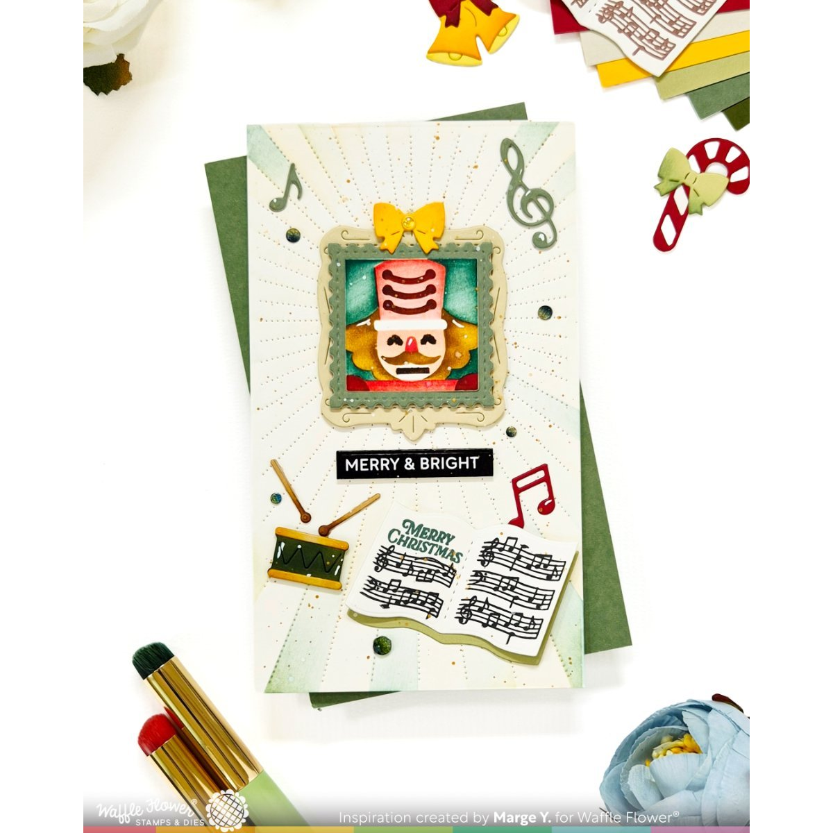 A handmade Christmas card featuring a nutcracker illustration, musical notes, sheet music, and the text 'MERRY & BRIGHT' and 'MERRY CHRISTMAS,' styled on a white background with decorative craft props.