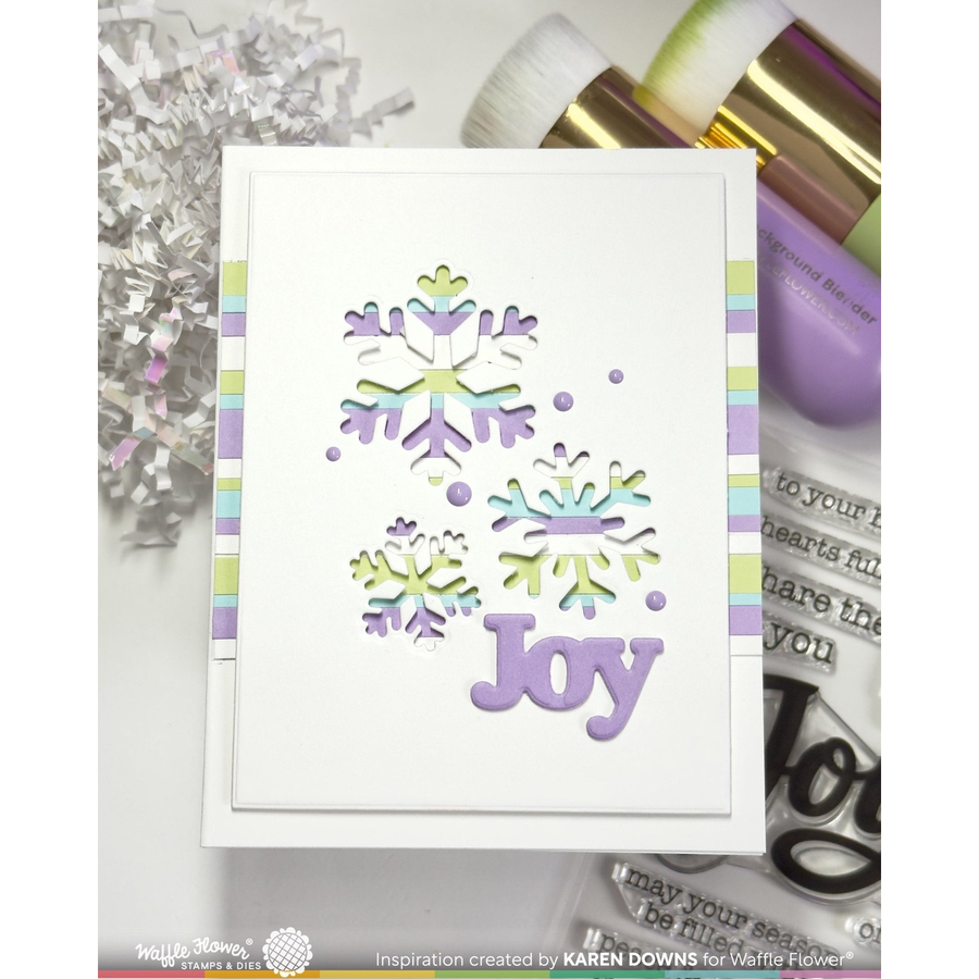 A handmade greeting card featuring layered white snowflakes and the word 'Joy' in lavender, shown with craft tools and silver tinsel in a styled flat-lay arrangement.