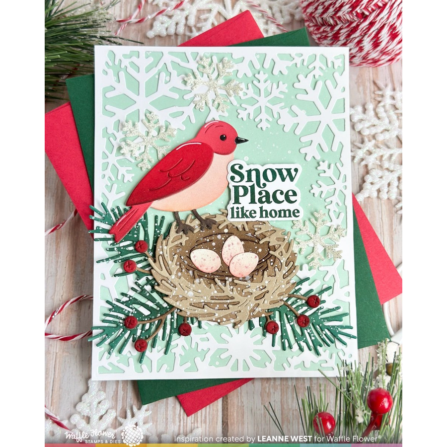 A handmade Christmas greeting card featuring a red bird perched above a nest with three eggs, surrounded by green pine branches, red berries, and a snowflake-patterned background. The card reads 'Snow Place like home' and is styled on top of red and green layered cardstock with holiday decorations around it.