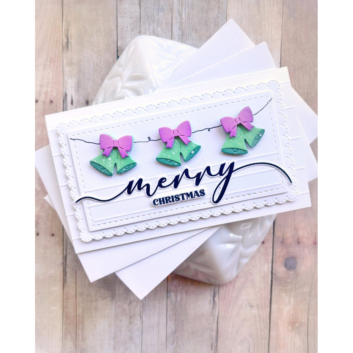 A handmade Christmas greeting card with three green bells and pink bows on a string, above the words 'merry CHRISTMAS' in black script and block lettering, displayed on top of other white cards over a light wood background.