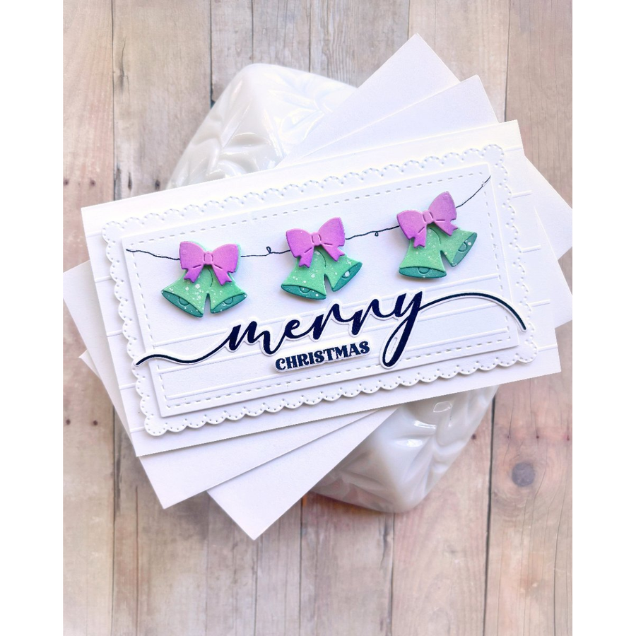 A handmade Christmas greeting card with three green bells and pink bows on a string, above the words 'merry CHRISTMAS' in black script and block lettering, displayed on top of other white cards over a light wood background.