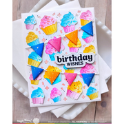 A handmade birthday card featuring colorful cupcake illustrations, bunting, and triangles, with a central die-cut label reading 'birthday WISHES'. The card is displayed on white napkins over a light wood surface.