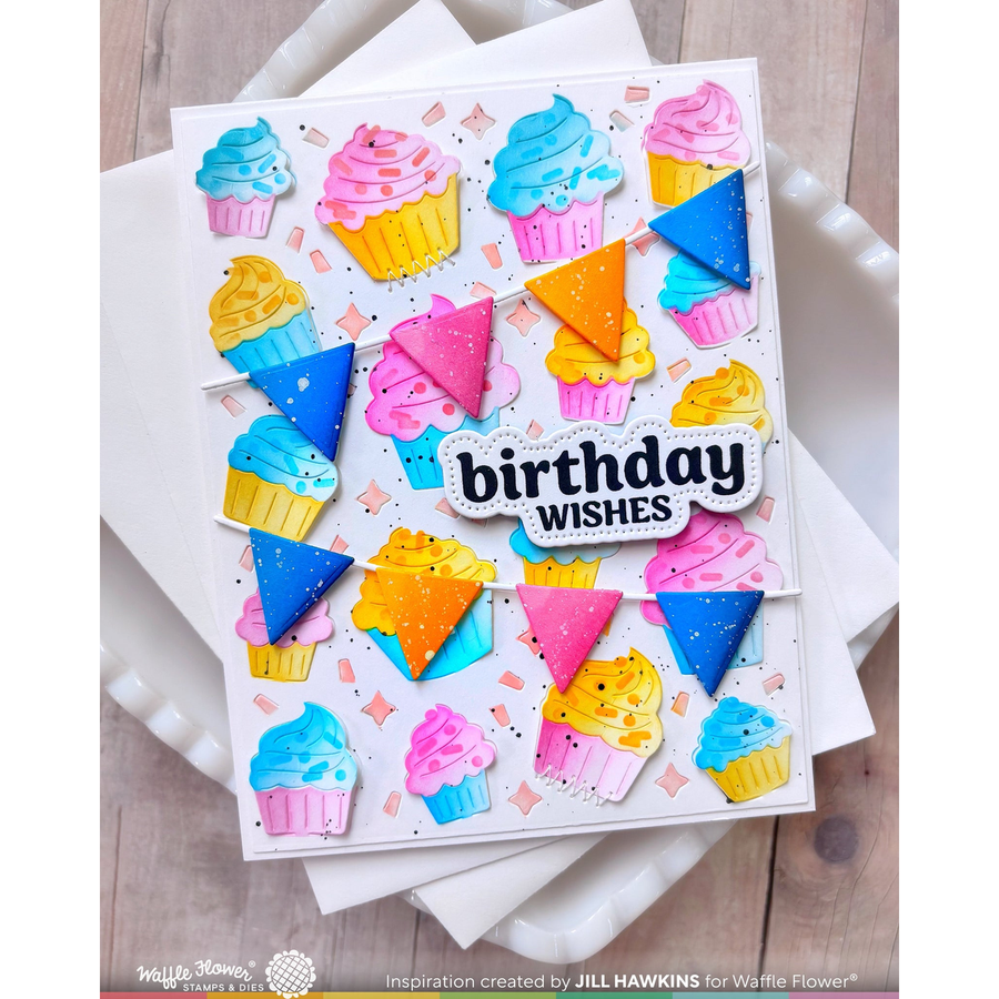 A handmade birthday card featuring colorful cupcake illustrations, bunting, and triangles, with a central die-cut label reading 'birthday WISHES'. The card is displayed on white napkins over a light wood surface.