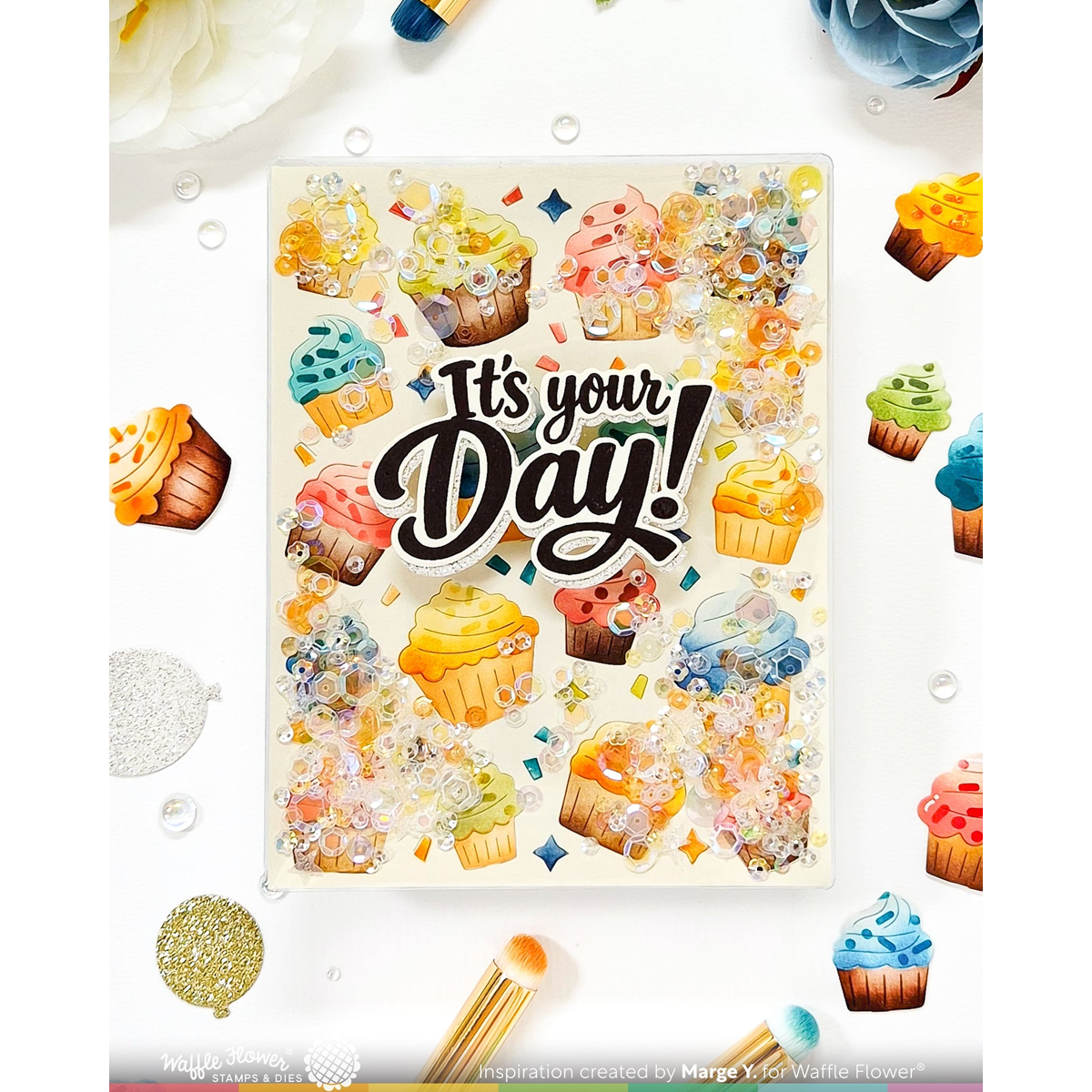 A square decorative card or art print with the phrase “It’s your Day!” in large black script, surrounded by colorful illustrated cupcakes, confetti, and glitter-like splashes on a white background. The card is photographed in a styled flat-lay with flowers, balloons, and paintbrushes around it.