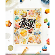 A square decorative card or art print with the phrase “It’s your Day!” in large black script, surrounded by colorful illustrated cupcakes, confetti, and glitter-like splashes on a white background. The card is photographed in a styled flat-lay with flowers, balloons, and paintbrushes around it.