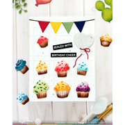 A handmade greeting card or print featuring colorful cupcake illustrations and bunting, with the text 'SEALED WITH BIRTHDAY CHEER' and a heart-shaped seal, shown on a light wood background with small decorative props.