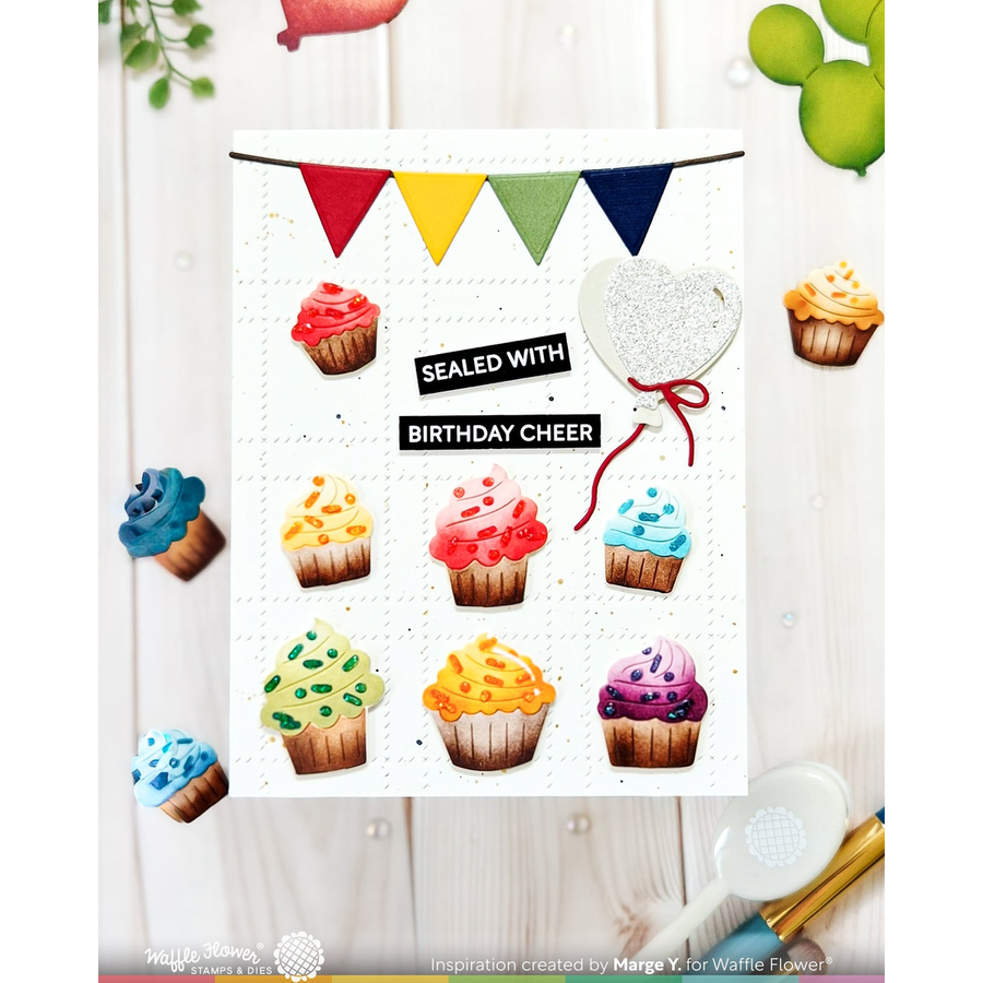 A handmade greeting card or print featuring colorful cupcake illustrations and bunting, with the text 'SEALED WITH BIRTHDAY CHEER' and a heart-shaped seal, shown on a light wood background with small decorative props.