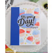 A handmade greeting card with a blue spine and a white front featuring the text 'It's your Day! enjoy it' and colorful cupcake illustrations, displayed on a white beaded plate.