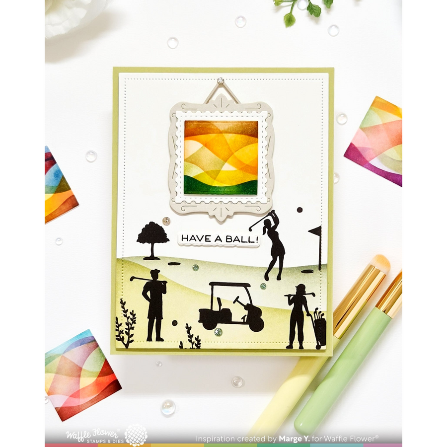 A handmade greeting card featuring a golf-themed design with black silhouette cutouts of golfers, a golf cart, and a framed abstract landscape in yellow, orange, and green. The card is displayed on a white background with craft pens and colorful paper accents.