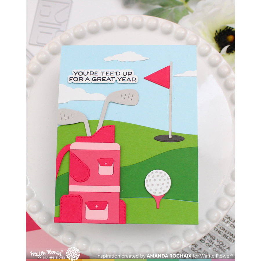 A handmade layered paper card featuring a golf course scene with a pink golf bag, golf clubs, a golf ball, and a flag, with the text 'YOU'RE TEE'D UP FOR A GREAT YEAR'. The card is displayed on a white beaded plate.