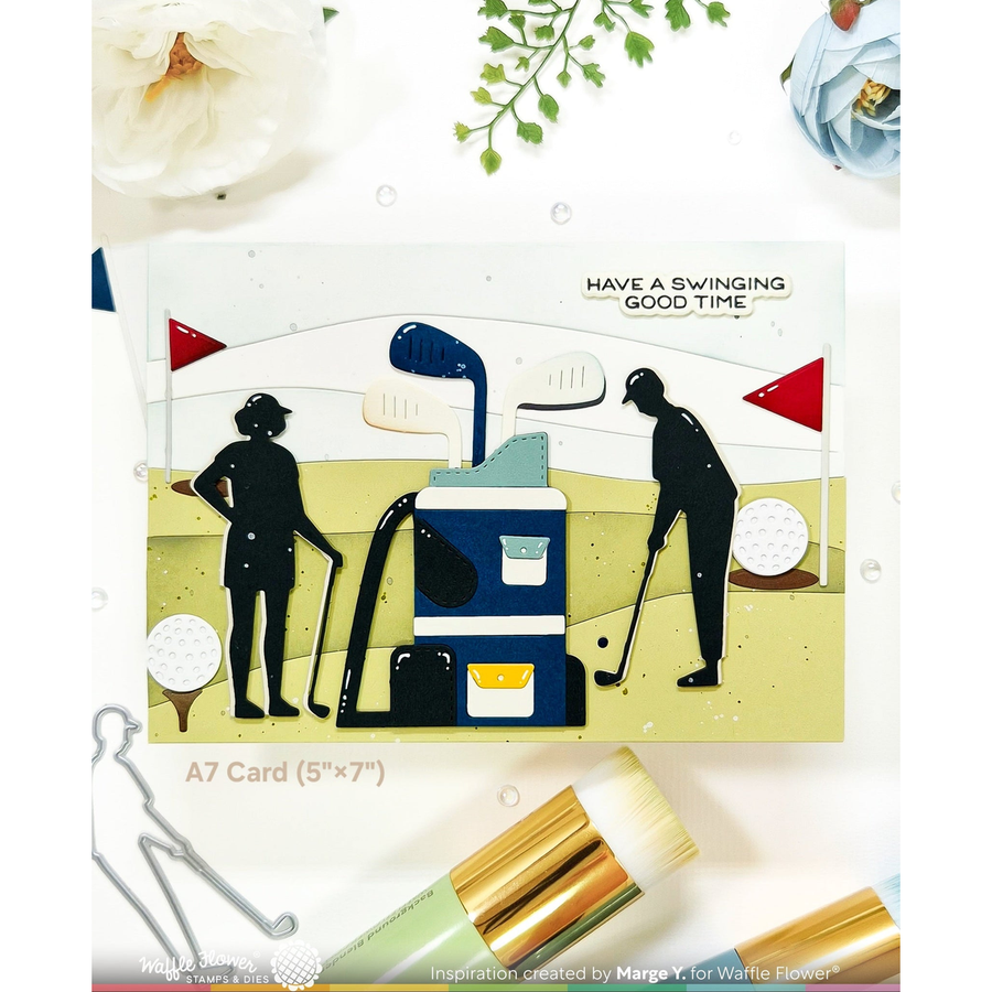 A die-cut A7 card featuring a golf-themed illustration with two black silhouettes of golfers, a blue and yellow golf cart, red flags, golf balls, and the text 'HAVE A SWINGING GOOD TIME.' The card is displayed on a white background with decorative flowers, paintbrushes, and small bubbles around it.