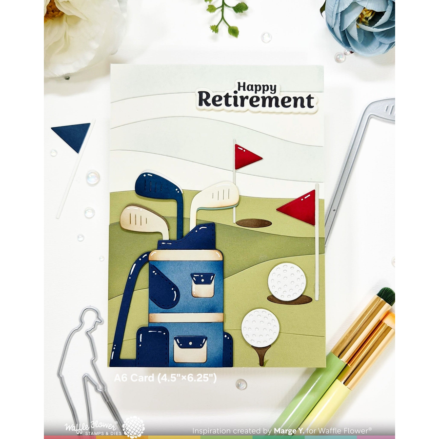 A handmade A6 card featuring a golf-themed retirement design with the words 'Happy Retirement' at the top, shown on a white flat-lay background with decorative flowers, metal cutout outlines, and golf props.