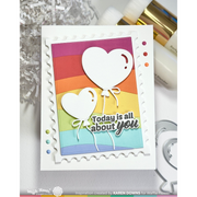 A handmade layered greeting card with scalloped white borders and two white heart balloons above the phrase 'Today is all about you.' The card features a rainbow striped background and is photographed on a light surface with craft supplies around it.