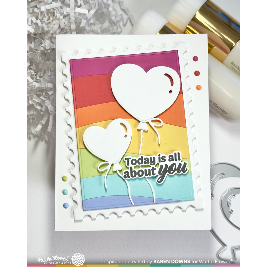A handmade layered greeting card with scalloped white borders and two white heart balloons above the phrase 'Today is all about you.' The card features a rainbow striped background and is photographed on a light surface with craft supplies around it.