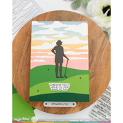 A handmade greeting card featuring a silhouette of a golfer on a green hill under a pastel sunset sky, displayed on a round wooden board with a small card label below it.