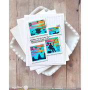 A handmade greeting card featuring postage stamp designs with camping and sailing themes, layered on top of white envelopes and a white dish over a rustic wood background. The card includes the text 'HOPE YOUR DAY IS SMOOTH SAILING'.