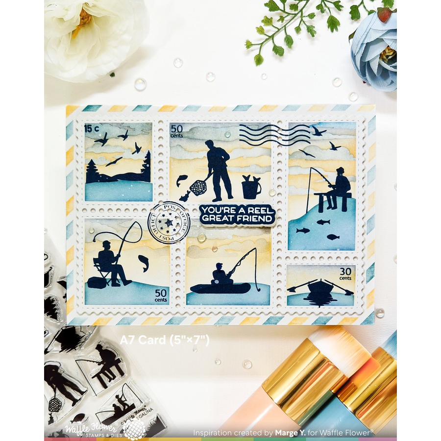 A rectangular A7 card featuring six postage-stamp-style fishing scenes with silhouettes, birds, fish, and the text 'YOU'RE A REEL GREAT FRIEND.' The card is photographed on a white surface with decorative flowers, greenery, and metallic gold-and-pink tools around it.