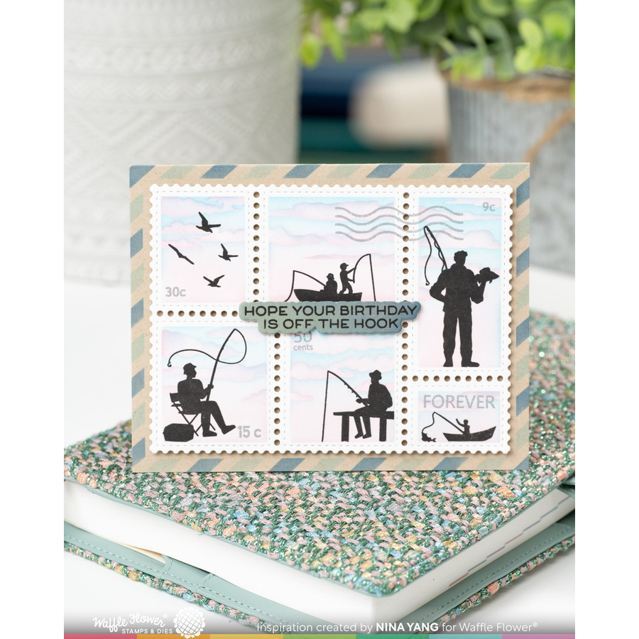 A rectangular fishing-themed birthday card displayed on top of a floral-patterned notebook. The card features six postage-stamp-like panels with black fishing silhouettes on a pale sky background and the text 'HOPE YOUR BIRTHDAY IS OFF THE HOOK' plus 'FISHING' and 'FOREVER'.