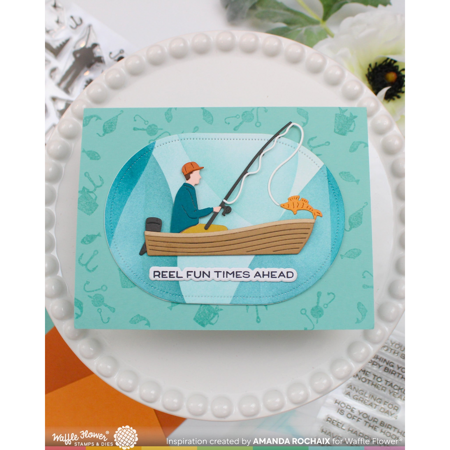 A handmade greeting card featuring a cartoon illustration of a man fishing in a wooden boat, displayed on a white scalloped plate. The card has a light blue background with subtle fishing-themed illustrations and the text 'REEL FUN TIMES AHEAD'.