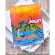 A handmade greeting card featuring a folk-art style illustration of a man fishing from a boat with a yellow fish jumping out, set against mountains and an orange sunset sky. The card is displayed on top of white envelopes on a light wood surface.