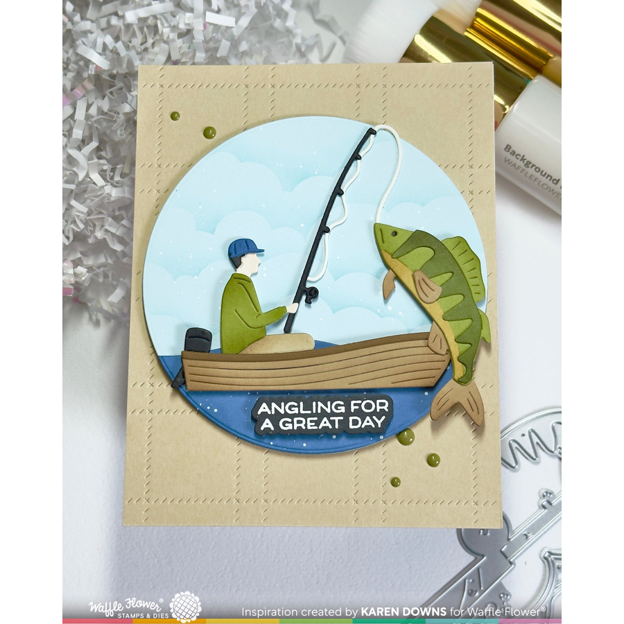 A handmade greeting card featuring a circular die-cut illustration of a person fishing in a boat and catching a large fish, with the text 'ANGLING FOR A GREAT DAY'. The card is displayed on a white surface with craft supplies in the background.