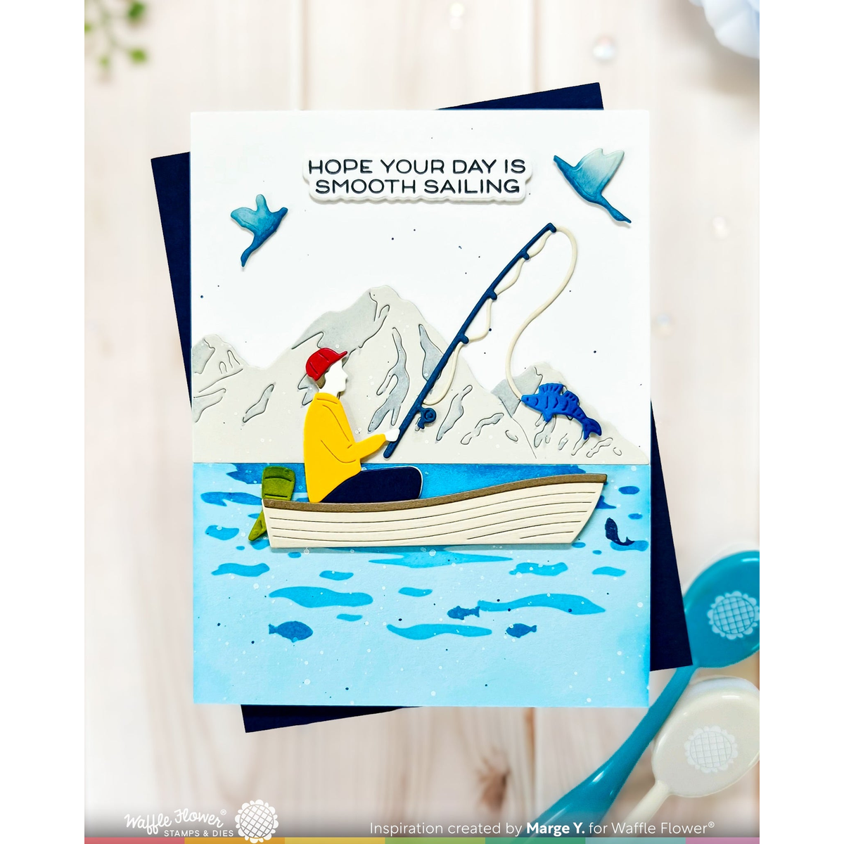 A handmade greeting card featuring a whimsical fishing scene with a person in a boat on blue water, with mountains and birds in the background. The card reads 'HOPE YOUR DAY IS SMOOTH SAILING.