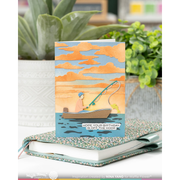 A greeting card featuring a handmade-style illustration of a person fishing in a boat under orange clouds, displayed on top of a floral-patterned notebook with potted plants in the background.