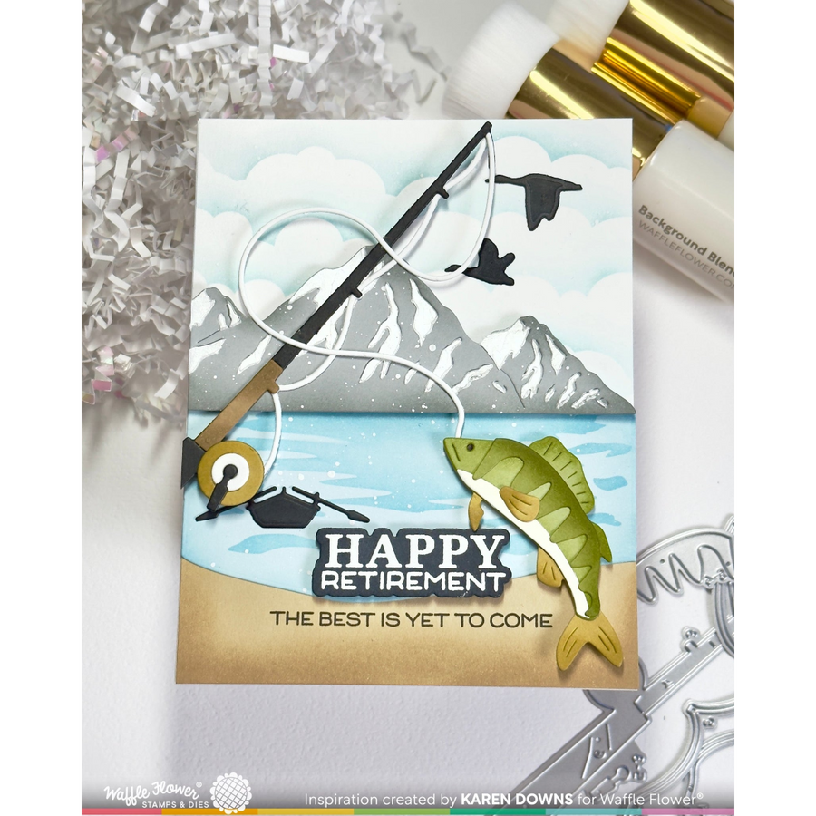 A handmade greeting card featuring a fishing theme with a fishing rod, fish, mountains, and the text 'HAPPY RETIREMENT THE BEST IS YET TO COME'. The card is displayed on a white surface with craft supplies in the background.