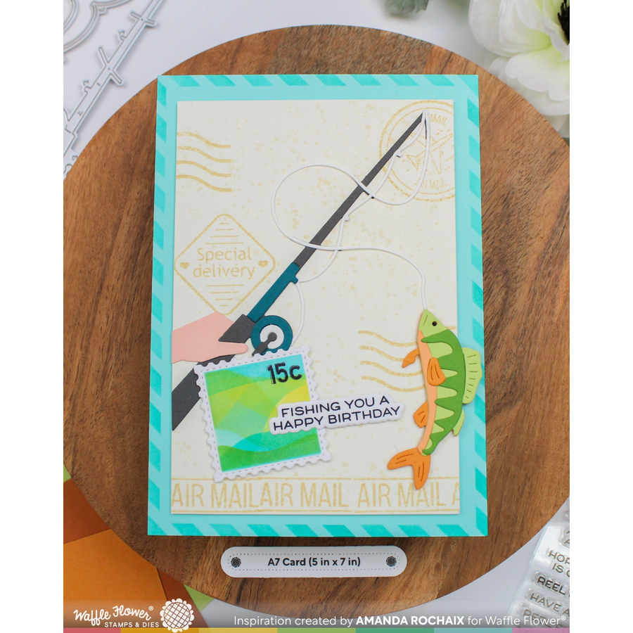 A handmade air mail style birthday greeting card with a fishing theme, displayed on a round wooden board. The card features a turquoise border, a fishing rod, a green and orange fish, and a stamp-like graphic with the text 'FISHING YOU A HAPPY BIRTHDAY'.