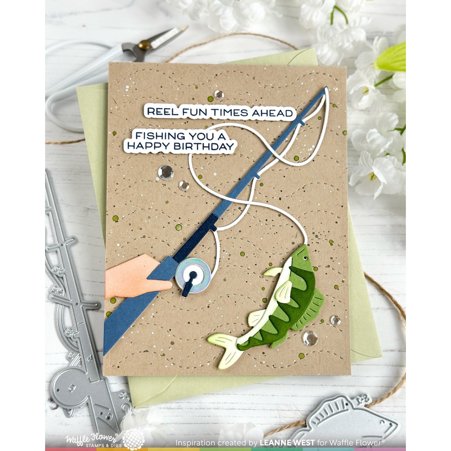 A handmade greeting card featuring a fishing theme, displayed on a white wooden surface with decorative props. The card has a speckled tan background, a blue and white fishing rod, a green and cream fish, and text that reads 'REEL FUN TIMES AHEAD' and 'FISHING YOU A HAPPY BIRTHDAY'.
