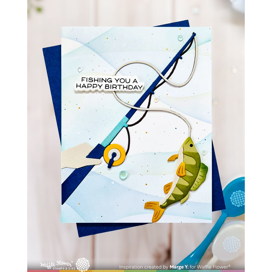 A handmade greeting card with a fishing theme, featuring a green and yellow fish on a hook, a blue fishing rod, and the text 'FISHING YOU A HAPPY BIRTHDAY'. The card is displayed on top of dark blue layered sheets, with a Waffle Flower logo and brand name visible at the bottom.