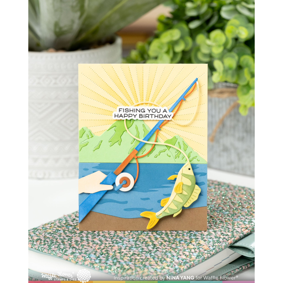 A handmade layered paper card with a fishing theme, featuring a blue lake, green hills, a yellow fish, and a fishing rod, displayed on a speckled notebook with a blurred plant background.