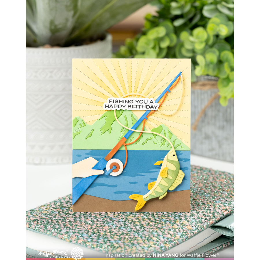 A handmade layered paper card with a fishing theme, featuring a blue lake, green hills, a yellow fish, and a fishing rod, displayed on a speckled notebook with a blurred plant background.