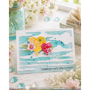 A handmade greeting card with a watercolor sky design, featuring two heart-shaped balloons in yellow and pink, decorated with small flowers. The card is displayed on a rustic white wooden surface surrounded by decorative props including a mason jar, flowers, and small blue glass beads.