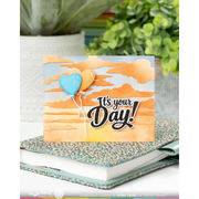 A handmade greeting card with a layered paper design of orange clouds and a blue heart-shaped balloon, reading 'It's your Day!' in black script. The card is displayed on top of a patterned notebook and a white book, with a blurred background of potted green plants.