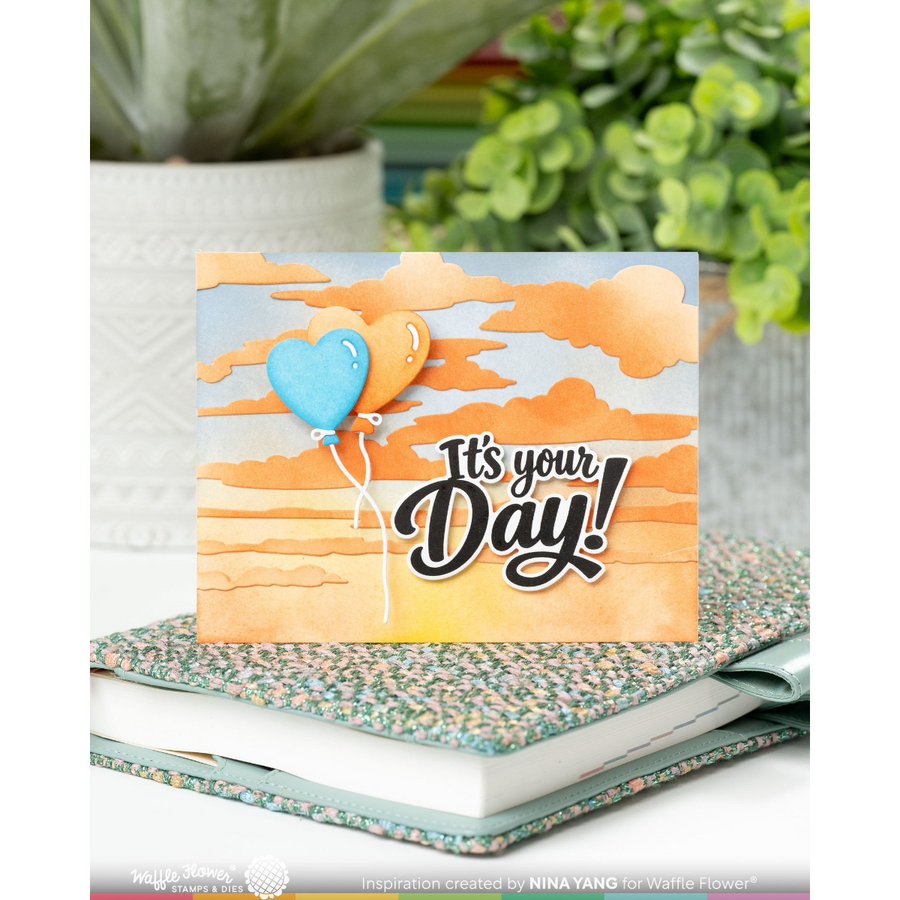 A handmade greeting card with a layered paper design of orange clouds and a blue heart-shaped balloon, reading 'It's your Day!' in black script. The card is displayed on top of a patterned notebook and a white book, with a blurred background of potted green plants.