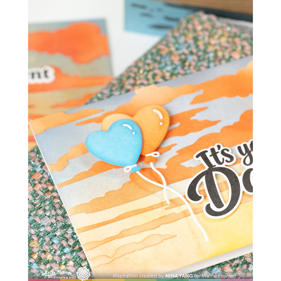 A close-up of a handmade greeting card featuring two heart-shaped balloons in light blue and orange against a watercolor sunset background, with partial text reading 'It's you Do' and glittery decorative paper underneath.
