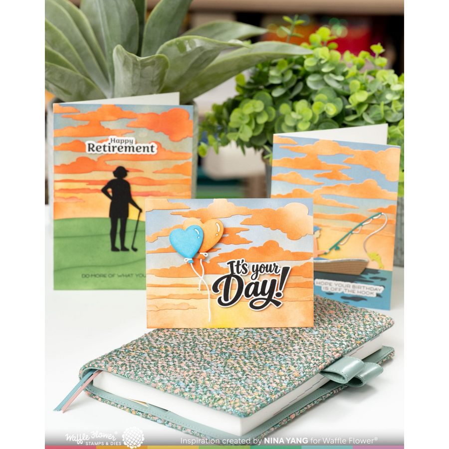 Three retirement-themed greeting cards displayed with a speckled notebook in the foreground, with potted green plants in the background. The cards feature warm sunset sky designs and text including 'Pension is Retirement' and 'It's your Day!'.