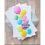 A handmade greeting card featuring colorful balloon illustrations and the text 'Celebrate you', displayed on top of white envelopes over a light wood surface with a white ceramic figurine in the background.