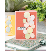 A handmade greeting card with a coral background and layered pastel balloon cutouts, standing on a speckled notebook with blurred green plants in the background. The card reads “HAPPY RETIREMENT” in a white label.