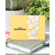 A yellow retirement greeting card with the words 'Happy Retirement' and a cluster of pastel balloon shapes, displayed on top of a patterned notebook with a blurred plant in the background.